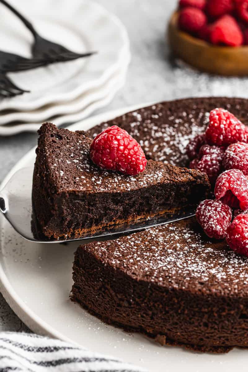 The best Flourless Chocolate Cake recipe dusted with powdered sugar and raspberries on top, with one piece being lifted to serve. 