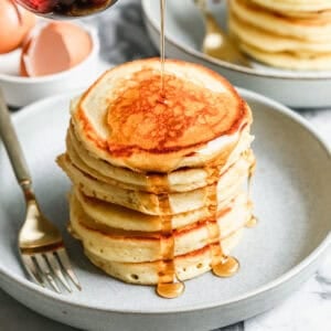 A stack of fluffy Buttermilk Pancakes on a plate with maple syrup being drizzled on top.