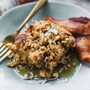 A piece of Hawaiian French Toast Casserole, sprinkled with coconut shavings and drizzled with syrup, ready to serve with a side of bacon.