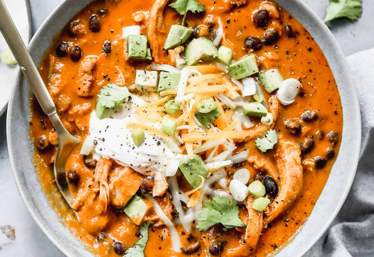 A bowl of a creamy Chicken Enchilada Soup recipe topped with chopped avocado, shredded cheese, and a scoop of sour cream.