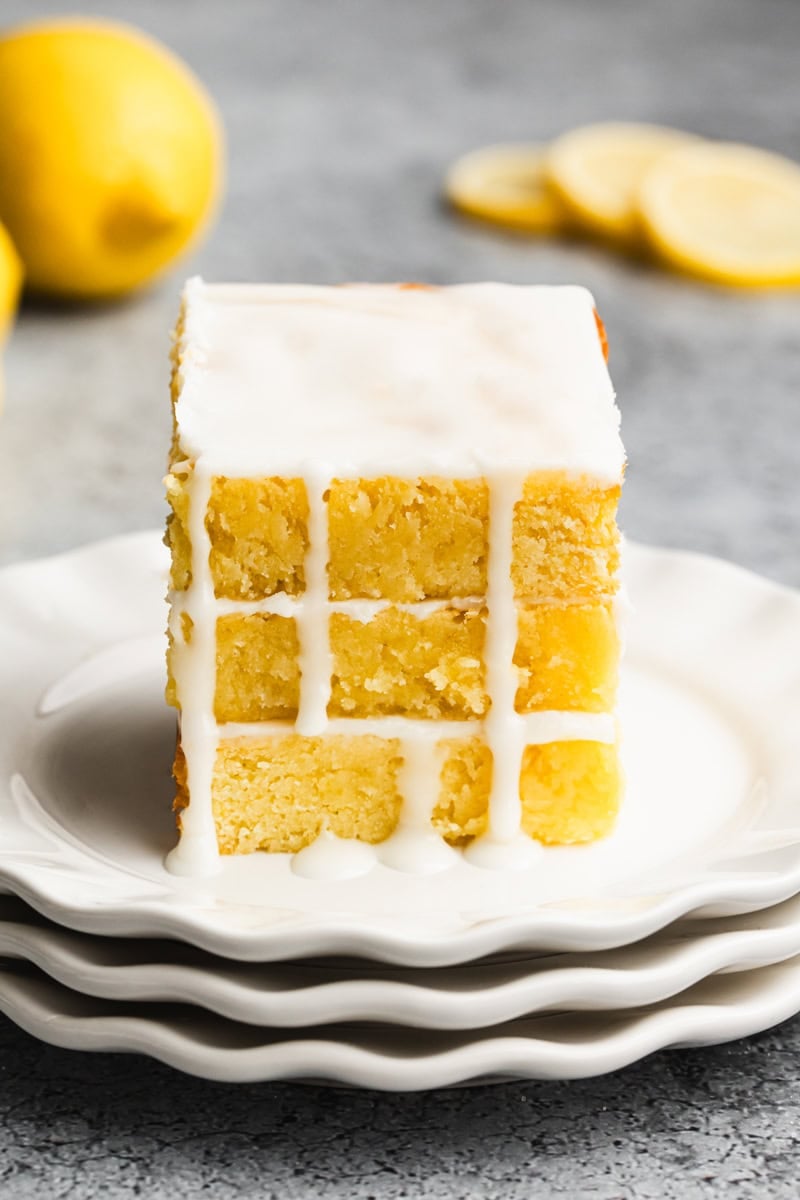 Three lemon brownies stacked on top of each other on a white plate with a simple glaze dripping down the sides.