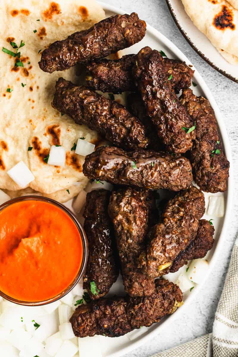 The best Cevapi recipe served on a plate with fresh pita bread, ajvar sauce, and sprinkled with chopped onion.