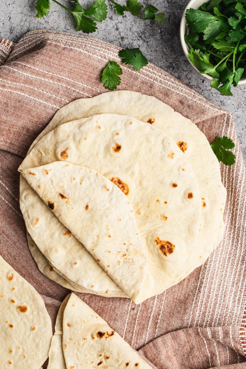 A stack of homemade flour tortillas on a clean kitchen cloth, ready to serve.