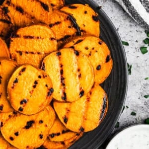 A plate filled with sliced Grilled Sweet Potatoes next to a cup of homemade dipping sauce.