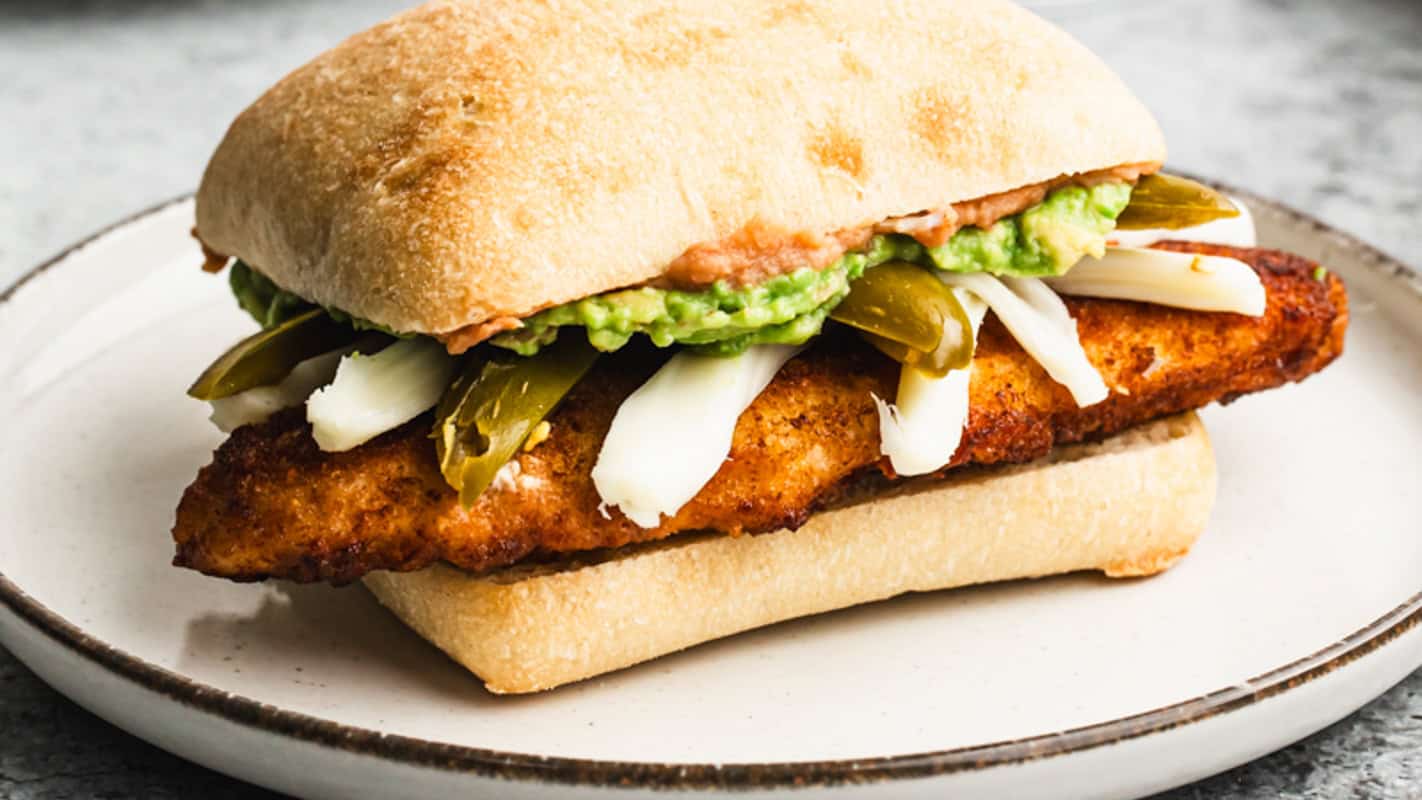 A Mexican Torta recipe with breaded, crispy chicken (chicken milanese), cheese, refried beans, guacamole, and pickled peppers on a white plate. 