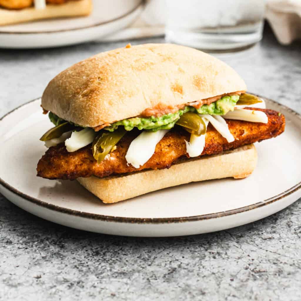 A Mexican Torta recipe with breaded, crispy chicken (chicken milanese), cheese, refried beans, guacamole, and pickled peppers on a white plate.