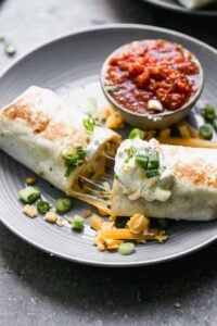 A breakfast burrito on a plate with salsa, sour cream and green onions on top.
