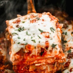 A serving of slow cooker lasagna being scooped out of a black crockpot with a large spoon, showing layers of noodles, sauce, and melted cheese.