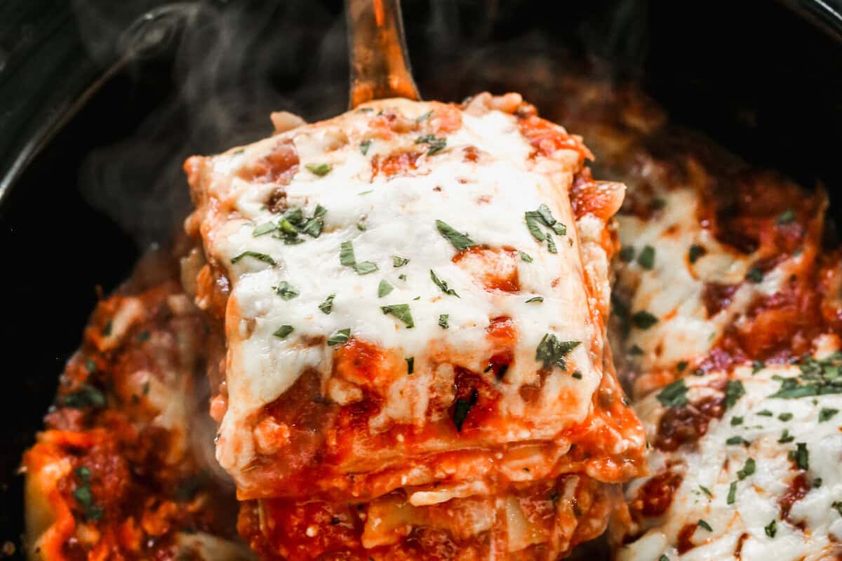 A serving of slow cooker lasagna being scooped out of a black crockpot with a large spoon, showing layers of noodles, sauce, and melted cheese.