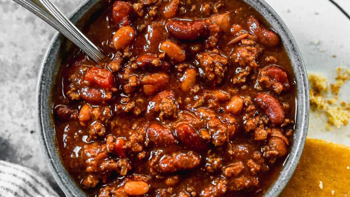 A bowl of homemade chili with ground beef, sausage and beans next to a piece of cornbread
