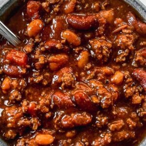 A bowl of homemade chili with ground beef, sausage and beans next to a piece of cornbread